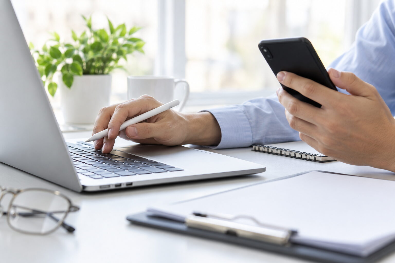 Hand mit Eingabestift an der Laptop-Tastatur, die andere Hand hält ein Smartphone, davor ein Klemmbrett mit leeren Blättern
